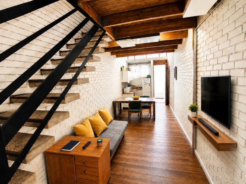 An open-plan living and kitchen area featuring exposed timber ceiling beams, painted brick walls, and warm timber flooring with a modern staircase.