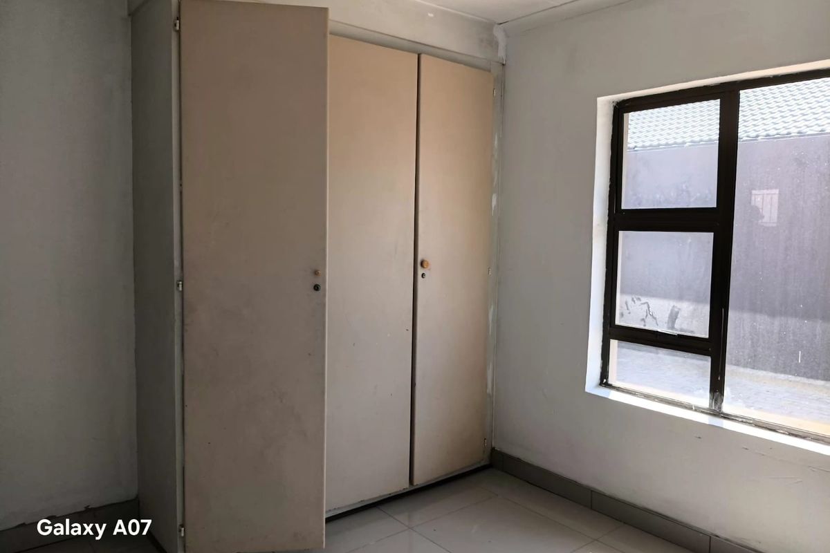 A vacant bedroom featuring tiled flooring, a dark aluminium window frame, and built-in cupboards with a dated finish.