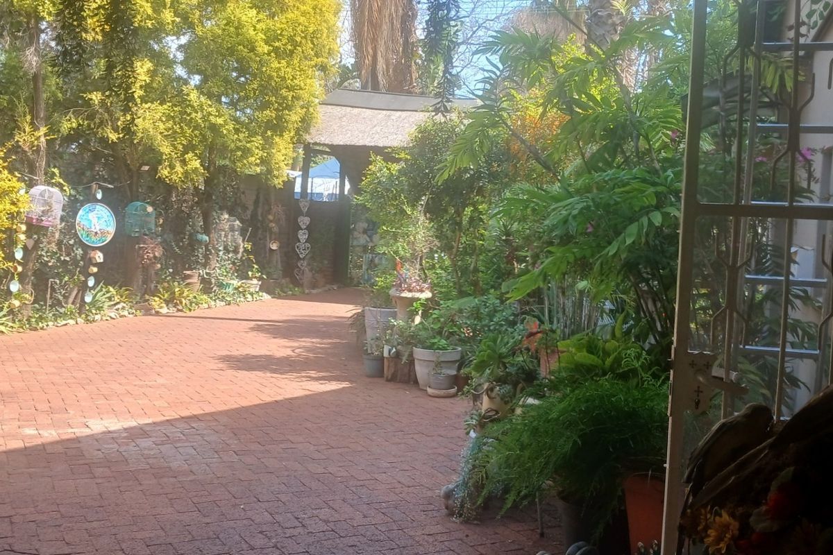 A rear garden area featuring brick paving, lush greenery, and a small thatched roof structure, viewed from a covered patio entrance.