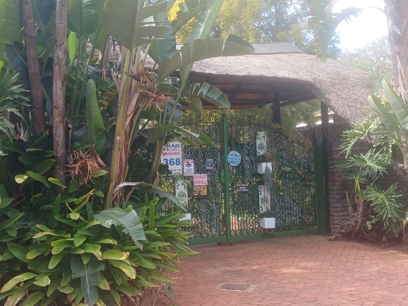 Front entrance featuring a thatched roof structure, brick driveway, and metal security gate surrounded by dense garden foliage.