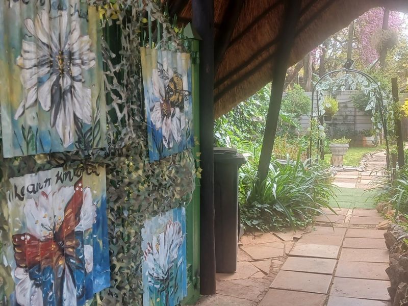 A lush garden entrance featuring a thatched roof walkway, stone paved path, and a decorative metal gate leading into a landscaped backyard area.