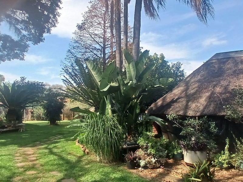 A lush, well-maintained garden featuring mature trees, a stone pathway, and a structure with a traditional thatched roof.