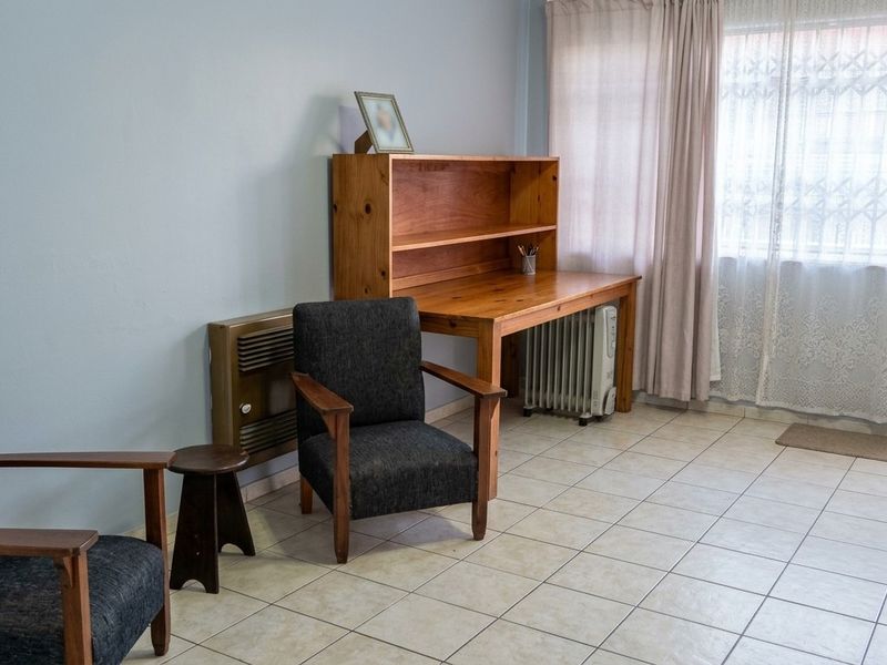 A living area featuring neutral tiled flooring, a wall-mounted heater, and a large window fitted with a security gate and curtains.