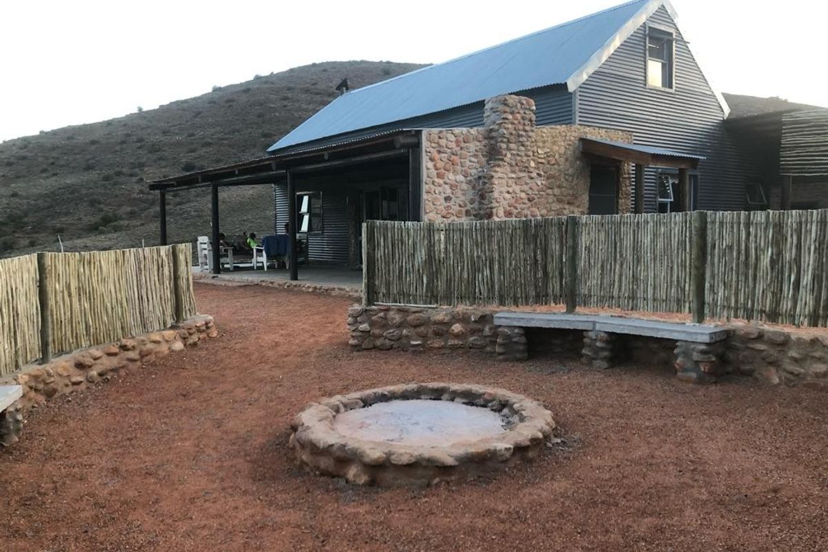 Rear exterior view featuring a corrugated metal roof house, covered patio, and a stone-built fire pit area with rustic timber fencing.
