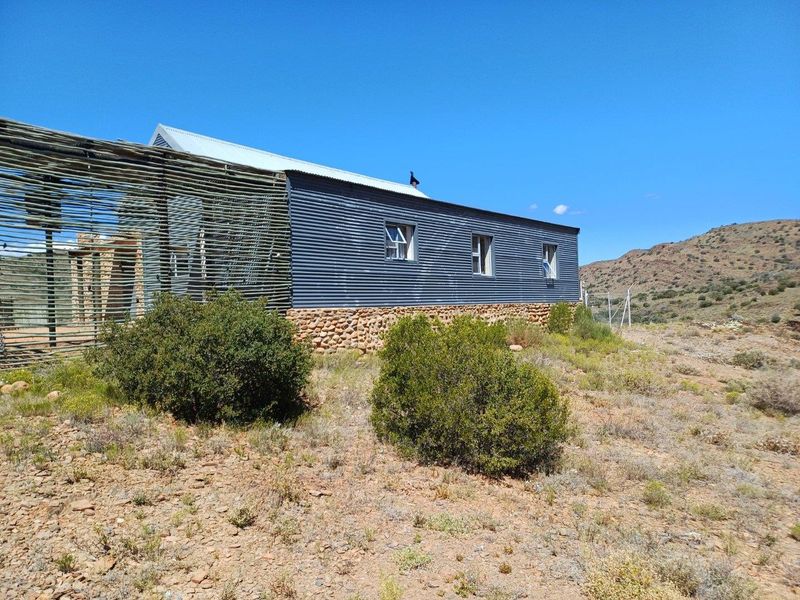 Rear view of a rural property featuring corrugated metal siding, a stone foundation, and a decorative timber slat screen set against a natural landscape.