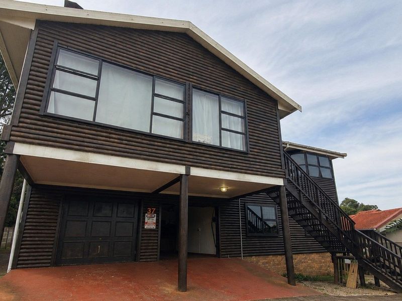 A two-story timber-clad house featuring a raised deck, external staircase, and ground-level garage access with a paved driveway.