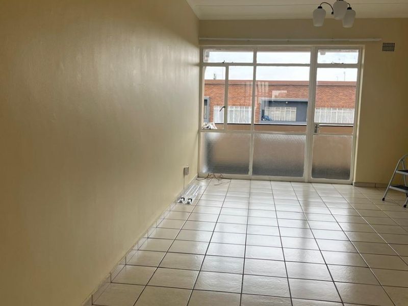 An empty, neutrally painted living room featuring tiled flooring and a large glass door providing natural light and access to the exterior.