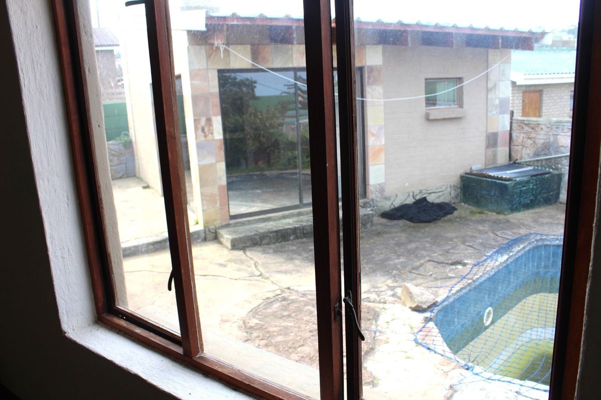 View from an interior window overlooking a rear garden area featuring a swimming pool with safety net, paved patio, and stone-clad exterior wall.