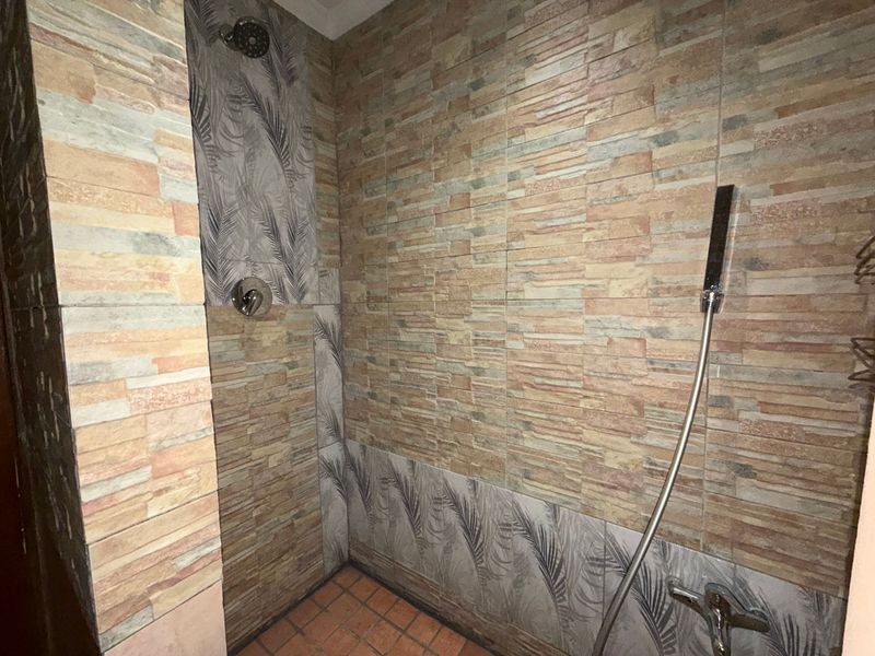 A shower enclosure featuring textured stone-look wall tiles and patterned accent tiles, equipped with both a fixed shower head and a handheld shower unit.