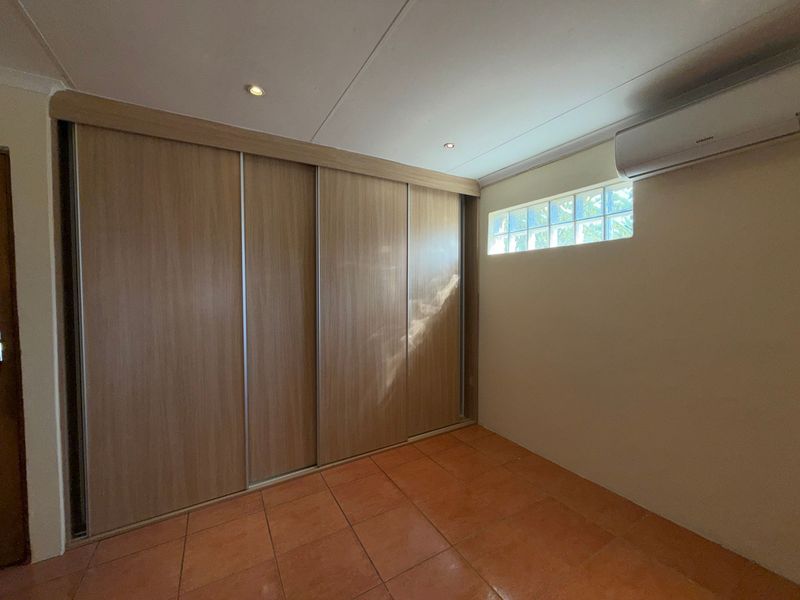 A clean, empty bedroom featuring built-in sliding door wardrobes, tiled flooring, a glass block window, and a wall-mounted air conditioning unit.