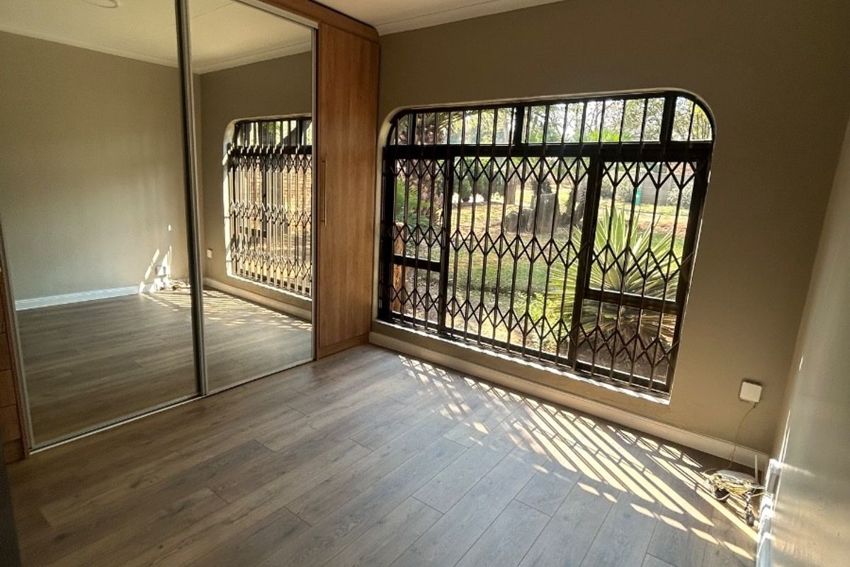 A bright, empty bedroom featuring modern laminate flooring, built-in cupboards with mirrored sliding doors, and a window fitted with a security gate.