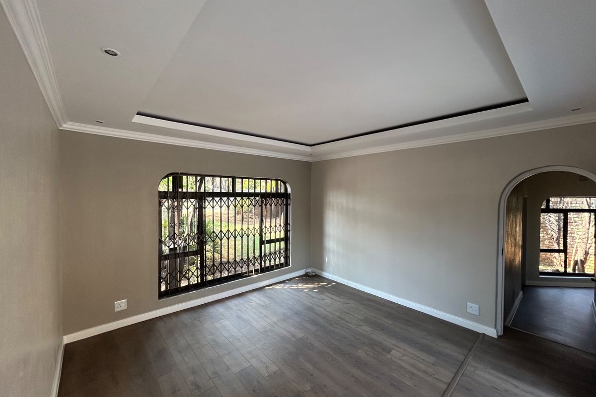 A bright, empty living room featuring modern laminate flooring, a recessed ceiling with coving, and a secure window with a metal security gate.