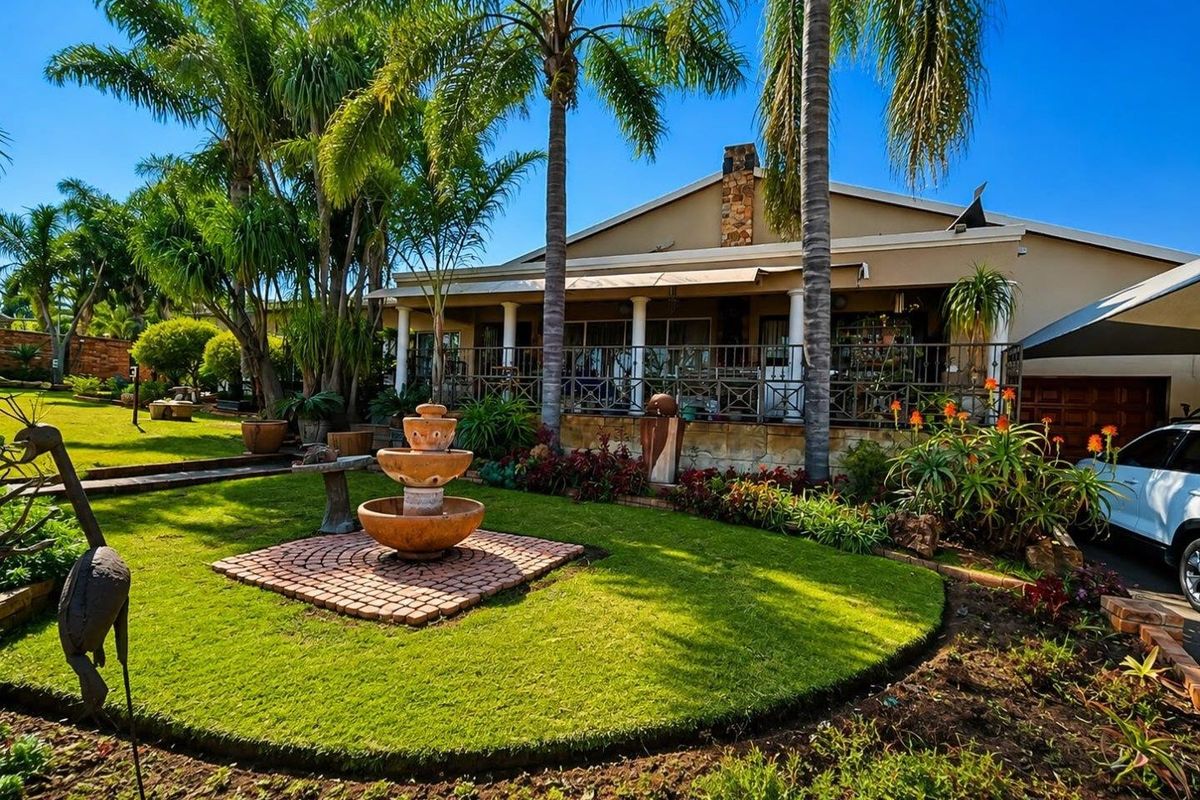 A well-maintained rear garden featuring a stone fountain, lush lawn, and a spacious covered patio with wrought iron railings and stone chimney detail.