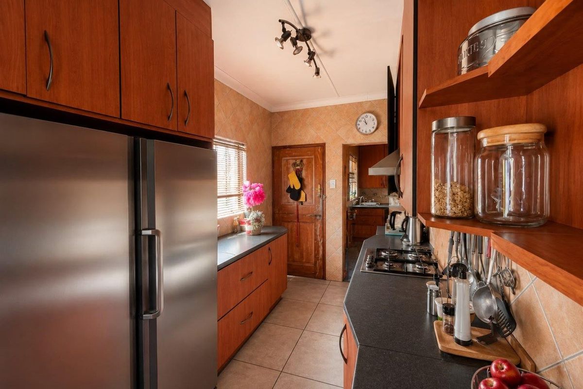 A functional kitchen featuring wood-finish cabinetry, dark countertops, a gas hob, and tiled finishes, with a view through to an adjacent scullery area.