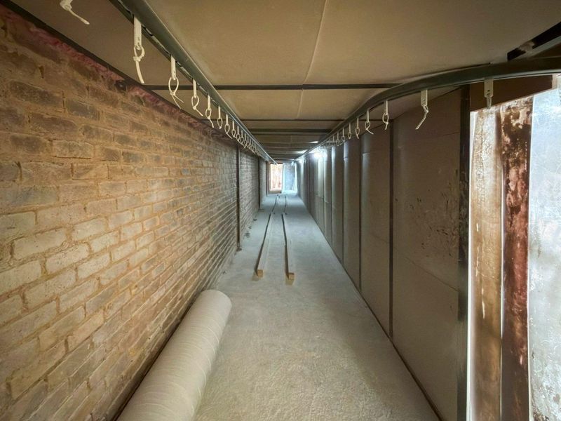 An industrial utility corridor featuring exposed brickwork, concrete flooring, and an overhead hanging rail system, suitable for storage or specialized industrial operations.