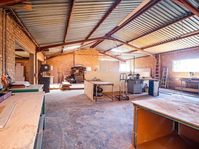 Spacious industrial workshop featuring exposed brick walls, high corrugated metal roofing with steel trusses, and durable concrete flooring, suitable for various manufacturing or storage uses.