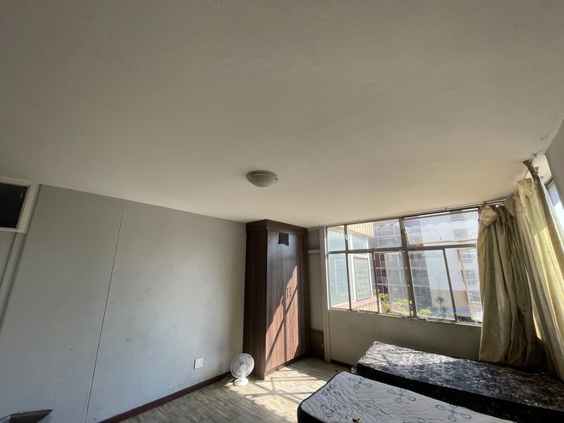 A bedroom featuring a built-in wardrobe, large windows providing natural light, and standard ceiling lighting. The room requires cosmetic updates to the wall finishes.