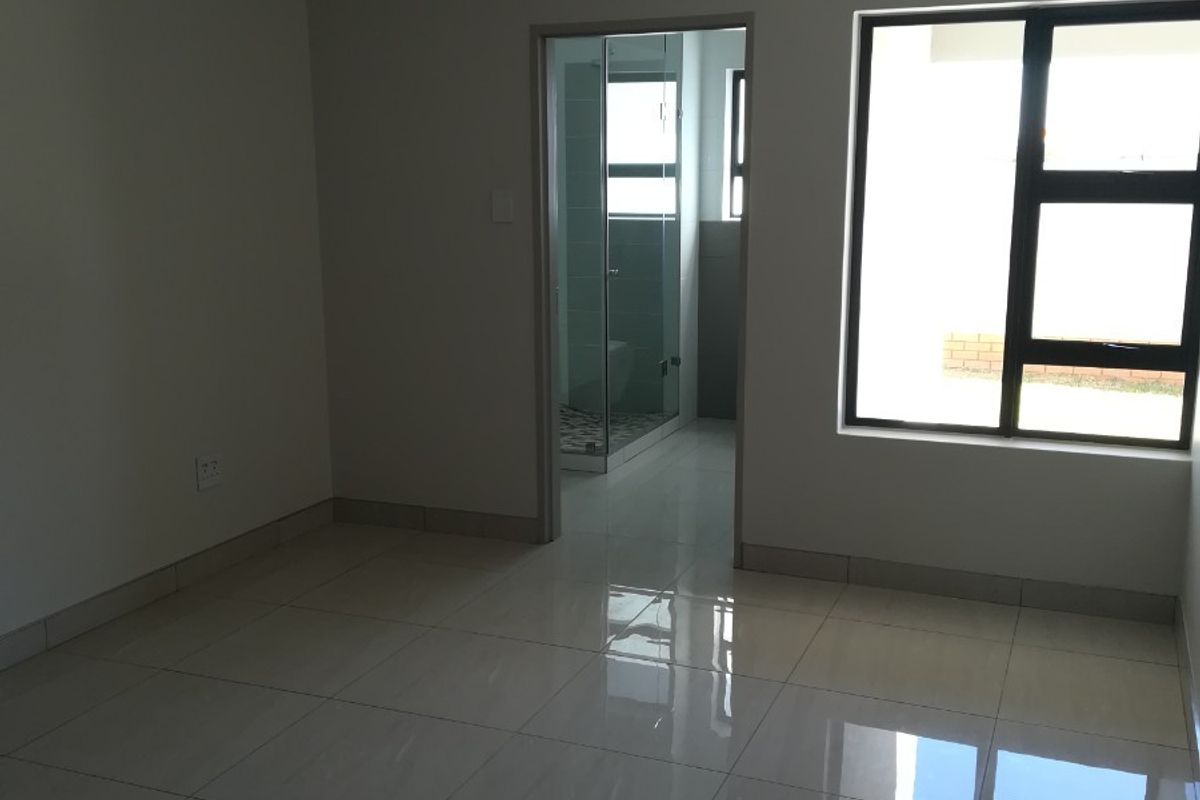 A bright, empty bedroom featuring modern tiled flooring, large aluminium-framed windows, and direct access to an en-suite bathroom.