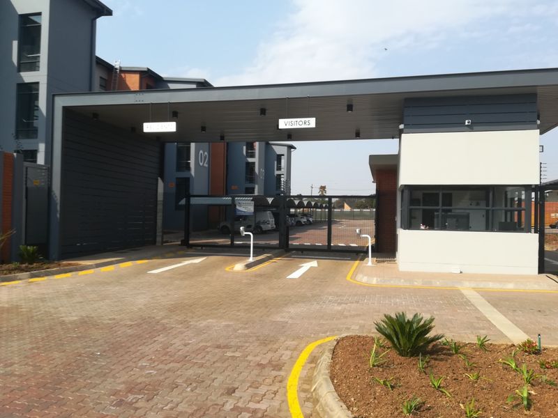 Modern secure residential complex entrance featuring a guard house, automated access control gates, and paved driveway with visitor parking signage.