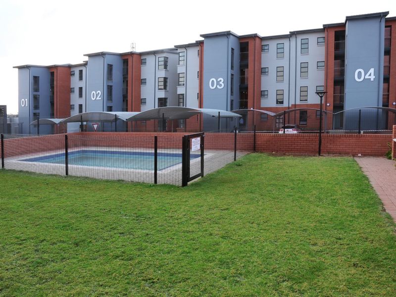 Communal pool area with a fenced swimming pool, brick boundary wall, paved walkway, and built-in braai facility, set within a modern townhouse complex.