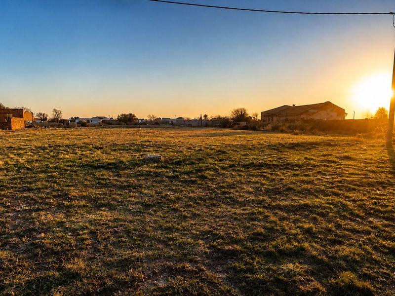 A wide-angle view of a vacant residential plot at sunset, featuring open grass, a utility pole, and surrounding boundary walls.