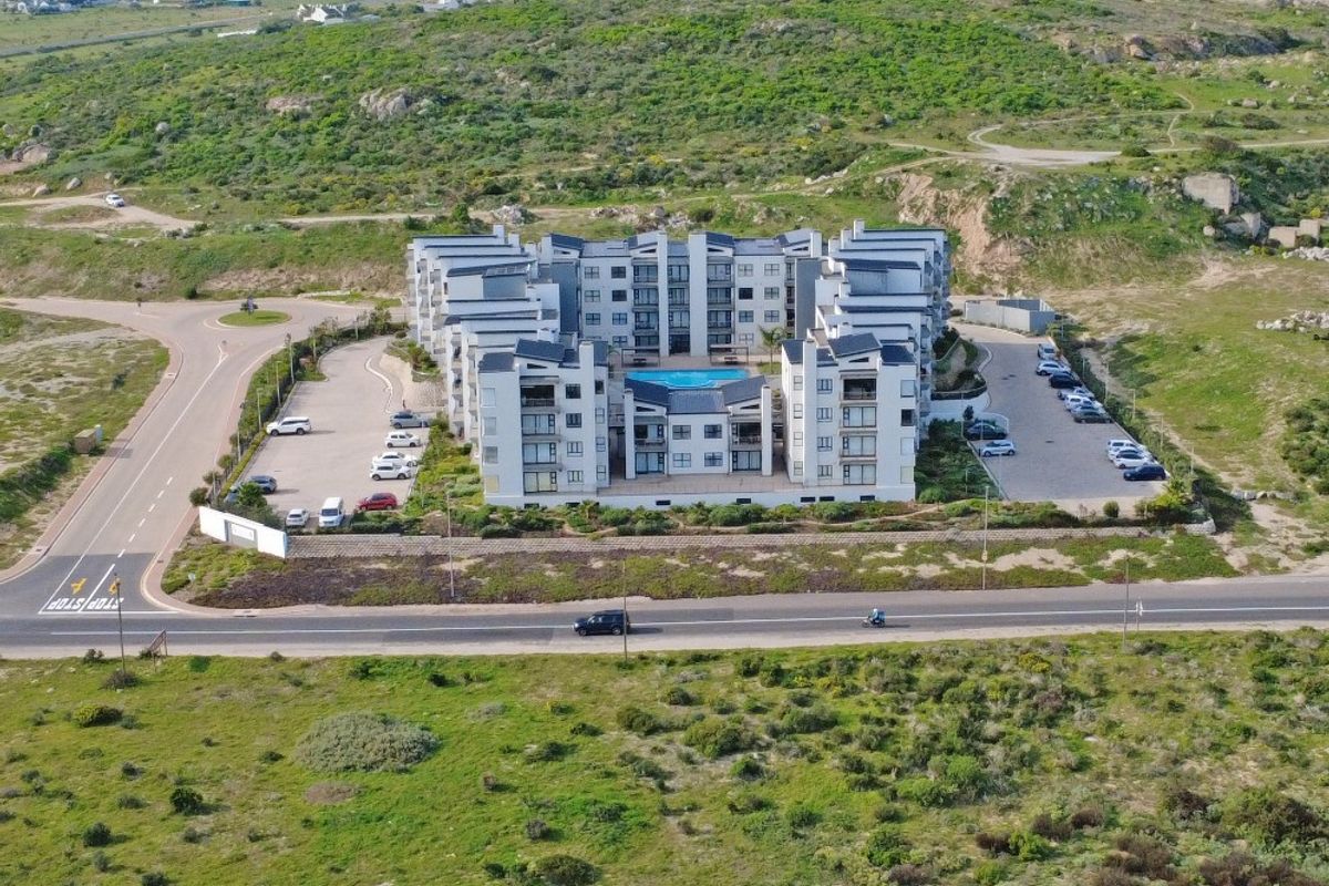 Aerial view of a modern multi-story apartment complex featuring a central swimming pool and surrounding paved parking areas in a coastal landscape.