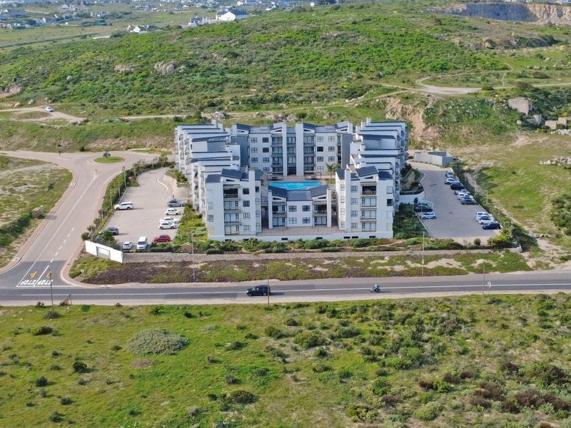 Aerial view of a modern multi-story apartment complex featuring a central swimming pool and surrounding paved parking areas in a coastal landscape.