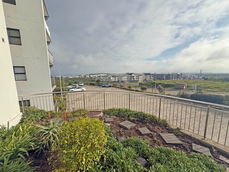 A balcony area featuring a metal balustrade and a small landscaped garden bed, overlooking a paved parking area and the surrounding neighborhood.