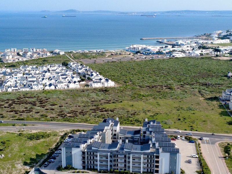 Aerial view of a modern apartment complex featuring rooftop solar panels, dedicated parking, and proximity to the coastline and surrounding residential areas.