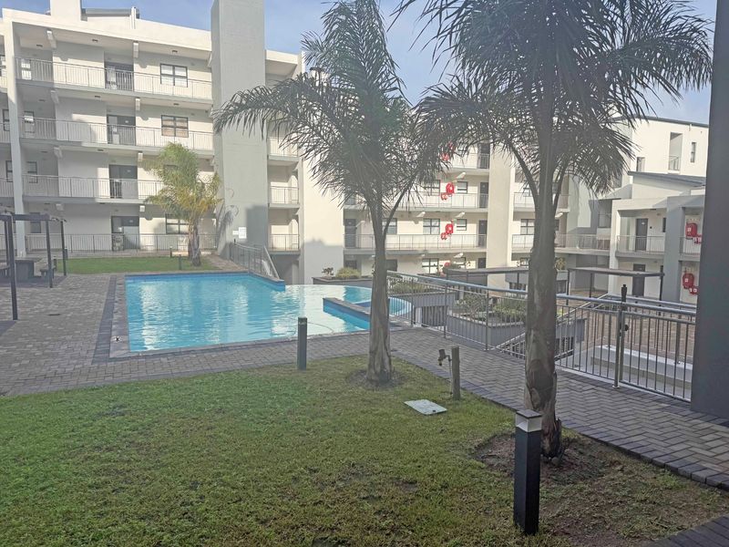 A well-maintained communal swimming pool area within an apartment complex, featuring paved walkways, metal safety railings, and landscaped garden elements.