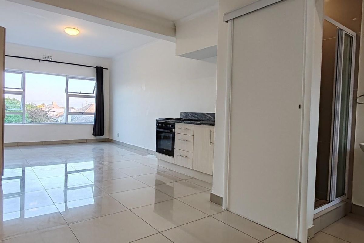 An open-plan apartment living area featuring tiled floors, a compact kitchen with built-in appliances, and an adjacent bathroom with a pedestal basin and shower.