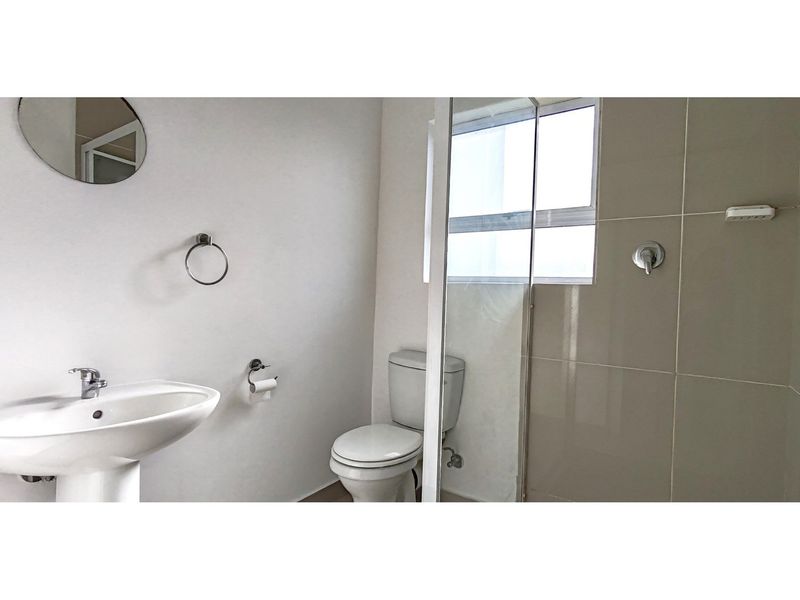 A clean, functional bathroom featuring a pedestal basin, toilet, and a glass-enclosed shower with neutral wall tiling.