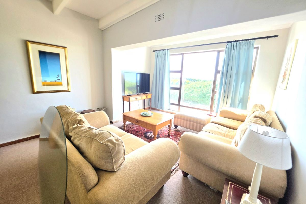 A comfortable living room featuring exposed ceiling beams, carpeted flooring, and a large window providing natural light and garden views.