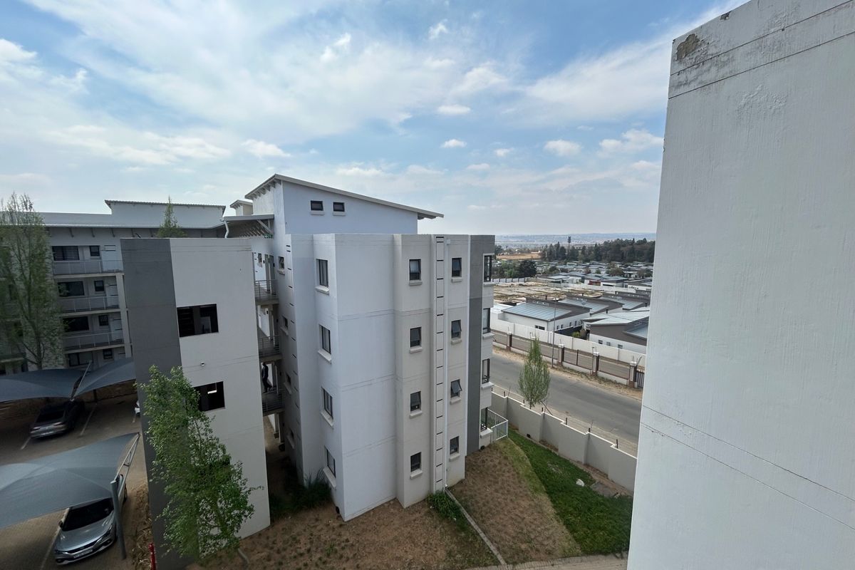 An elevated view of a modern apartment complex featuring multi-story residential blocks, paved parking with shade cloth carports, and surrounding perimeter fencing.