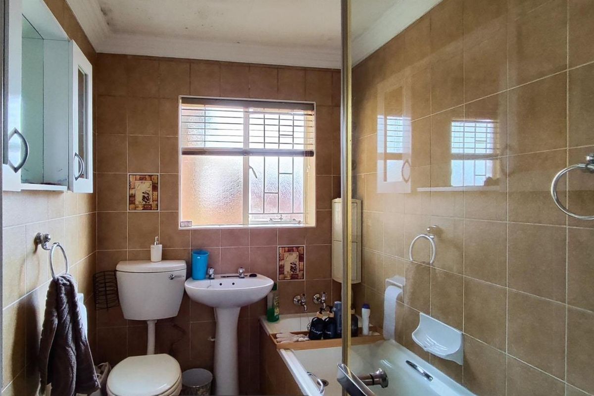 A compact bathroom featuring a pedestal basin, toilet, and bathtub with a shower screen, finished with dated brown wall tiles and a window.