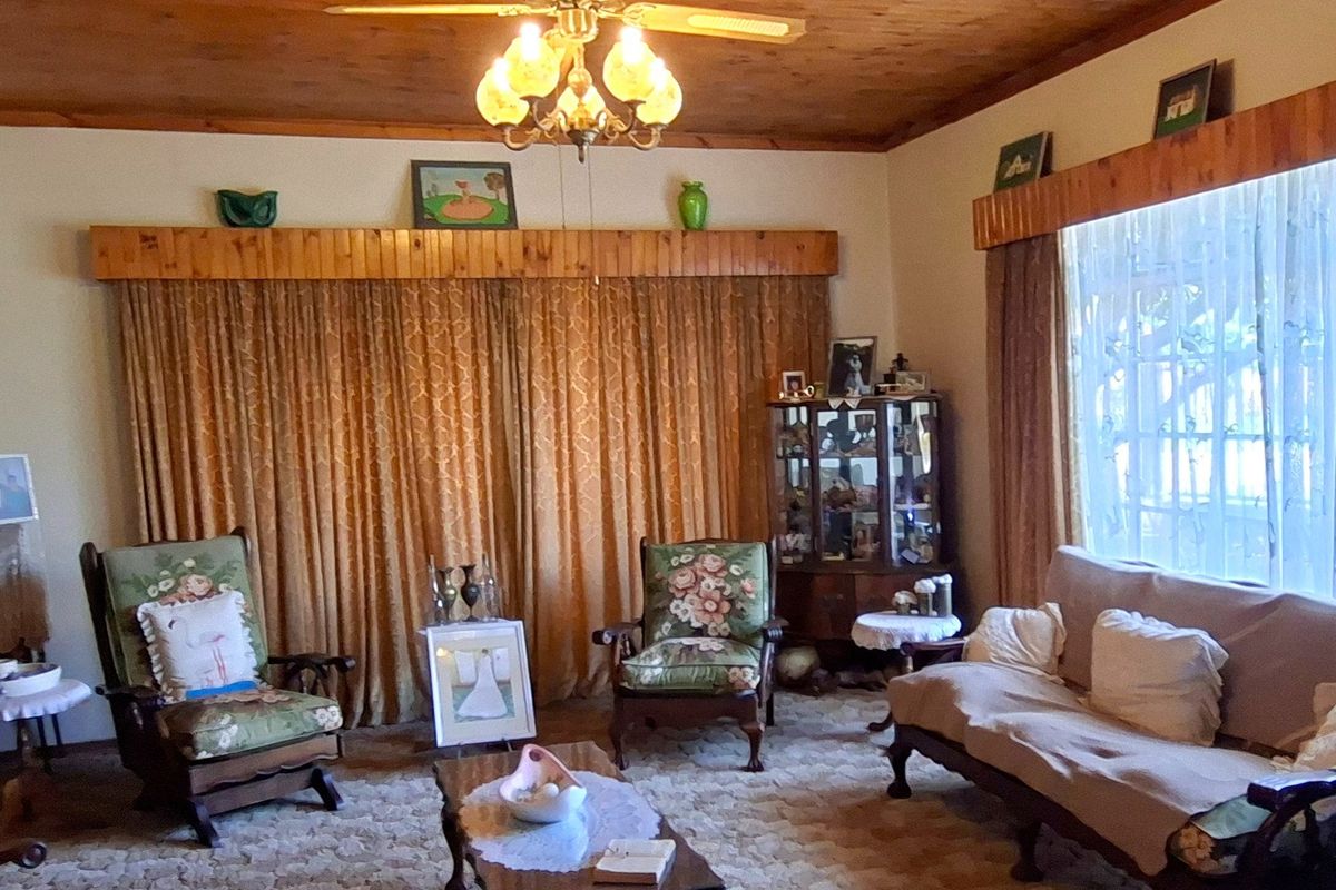 A traditional living room featuring a timber-paneled ceiling, ceiling fan, and large windows with timber pelmets, offering a dated but functional living space.