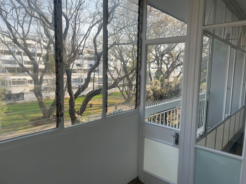 Enclosed balcony area featuring large windows with views of the surrounding complex grounds and mature trees.