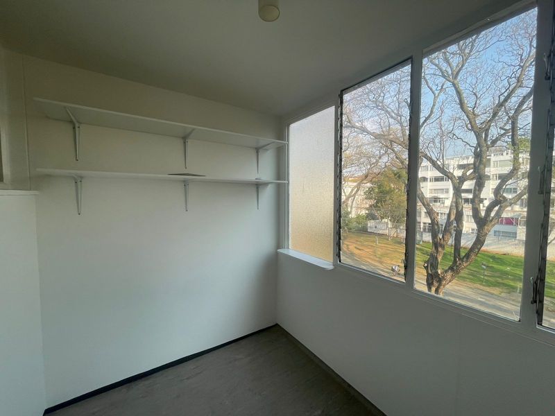 A bright, empty bedroom featuring laminate flooring, built-in wall shelving, and large louver windows with a pleasant neighborhood view.