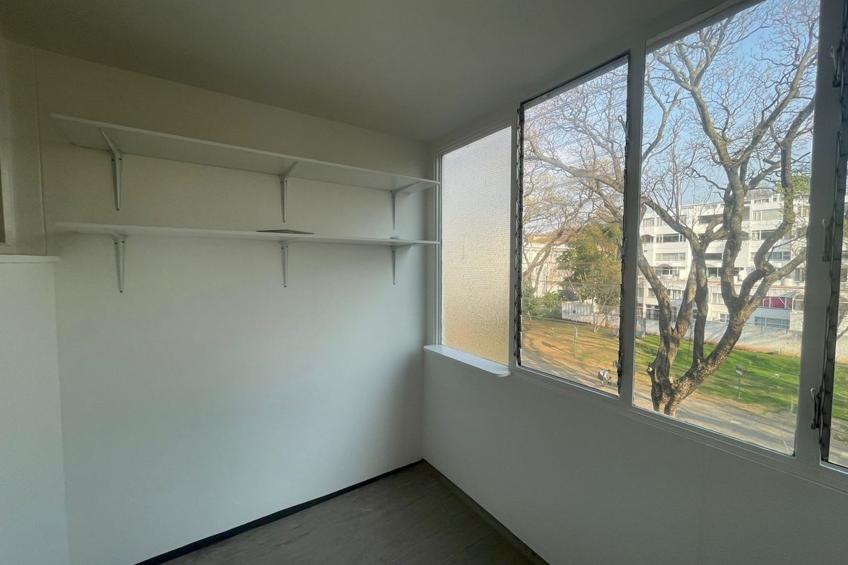A bright, empty bedroom featuring laminate flooring, built-in wall shelving, and large louver windows with a pleasant neighborhood view.