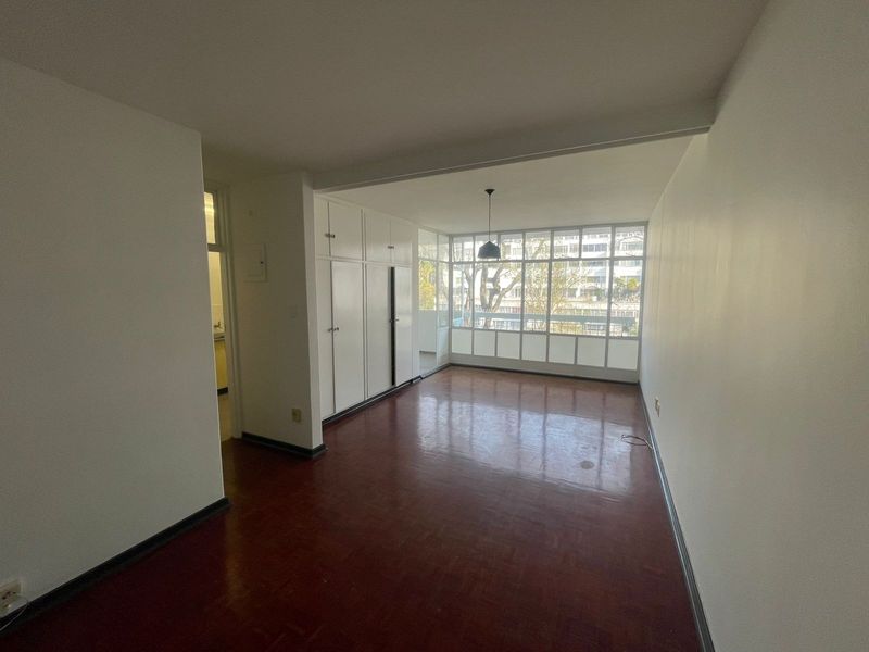 An empty living room featuring original parquet flooring, built-in storage cupboards, and a large window wall providing natural light and an urban outlook.