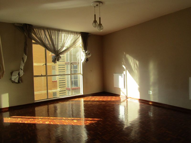 A bright, empty living room featuring polished parquet flooring, a large window with natural light, and a wall-mounted heater.