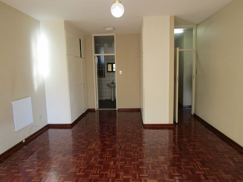 An empty bedroom featuring polished parquet flooring, built-in storage cupboards, and an adjoining en-suite bathroom area with a pedestal basin.