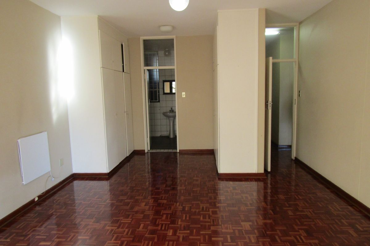 An empty bedroom featuring polished parquet flooring, built-in storage cupboards, and an adjoining en-suite bathroom area with a pedestal basin.