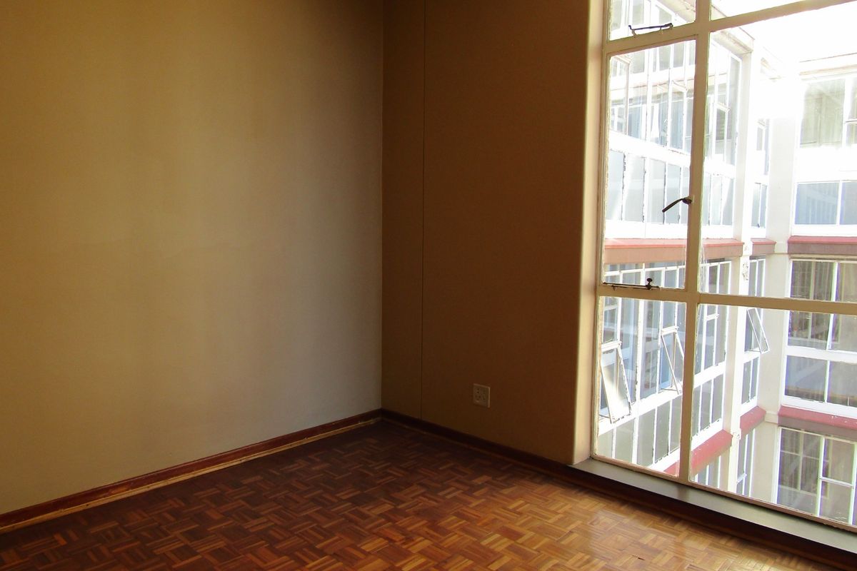 An empty bedroom featuring original parquet flooring and a large window overlooking the building's internal courtyard.