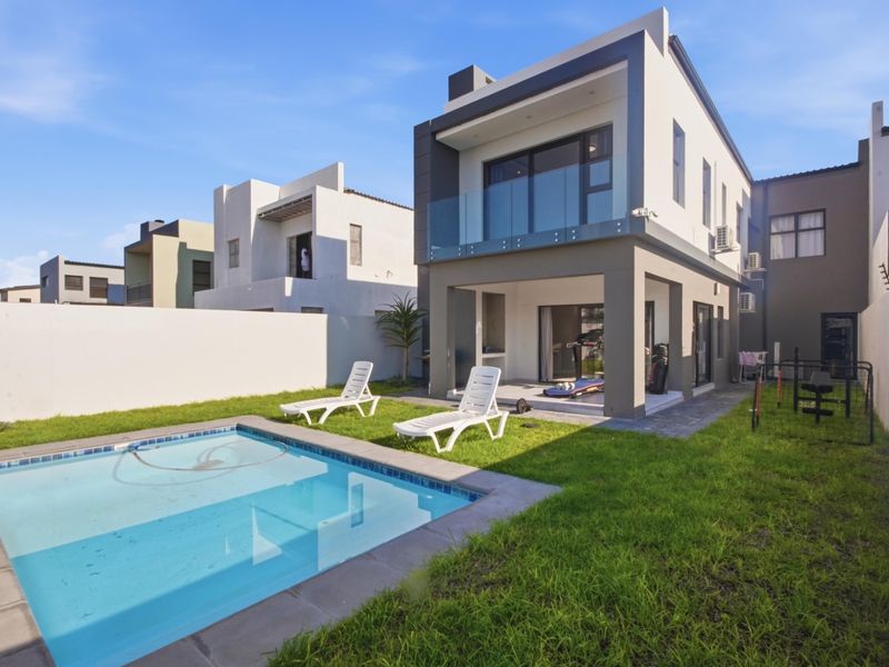Rear view of a modern double-story house featuring a swimming pool, landscaped garden, covered patio with built-in braai, and an upper-level balcony with glass balustrades.