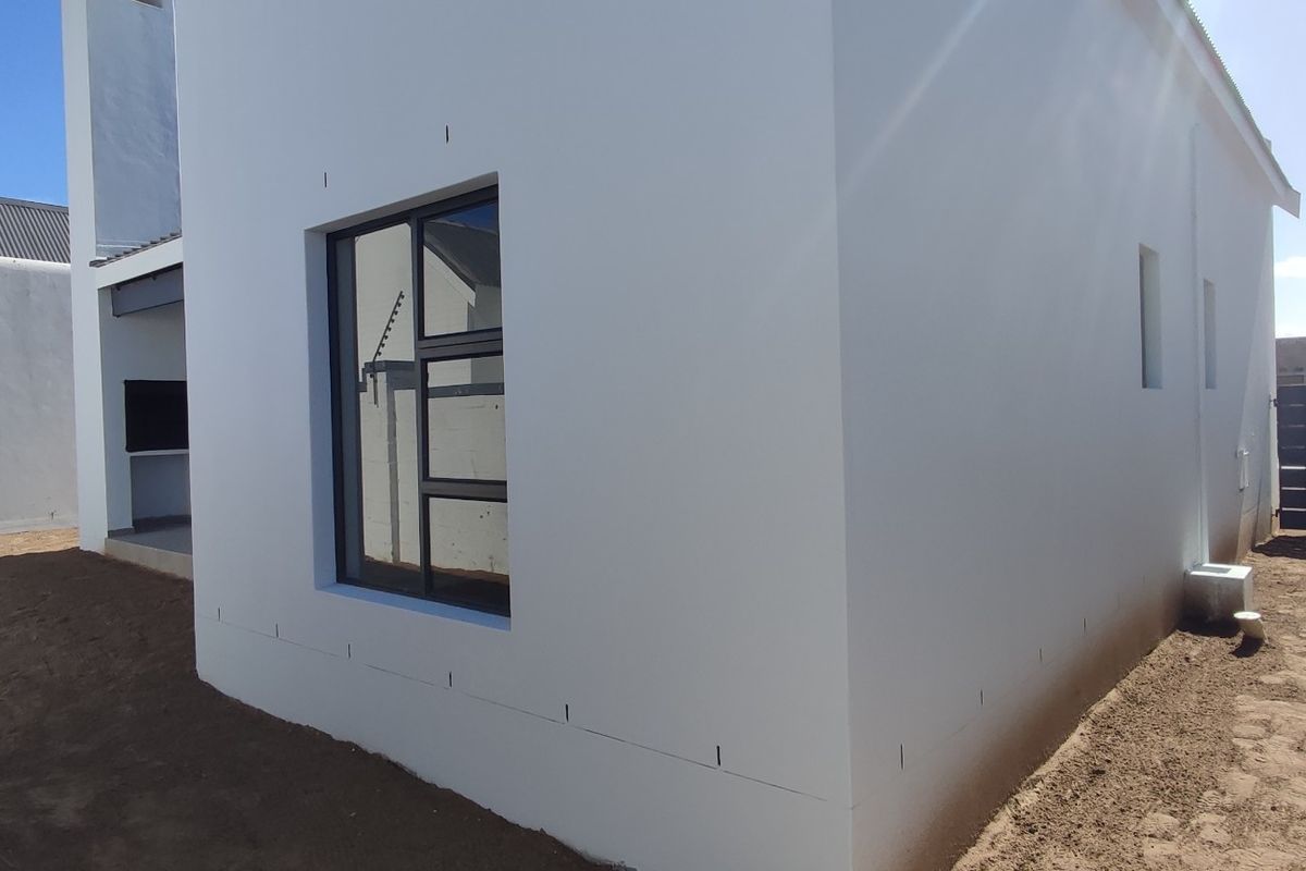 Rear view of a modern white house featuring a built-in braai area, aluminium window frames, and an unlandscaped garden space.