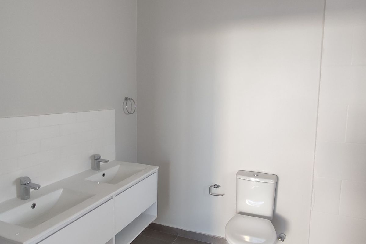 A modern, minimalist bathroom featuring a double vanity with dual basins, a toilet, and a tiled shower area with a wall-mounted shower head.