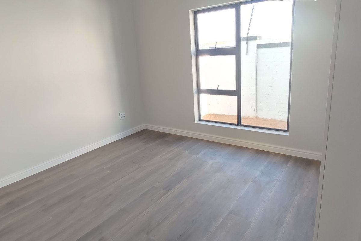 A clean, empty bedroom featuring modern laminate flooring, neutral walls, and a large aluminium-framed window providing natural light.