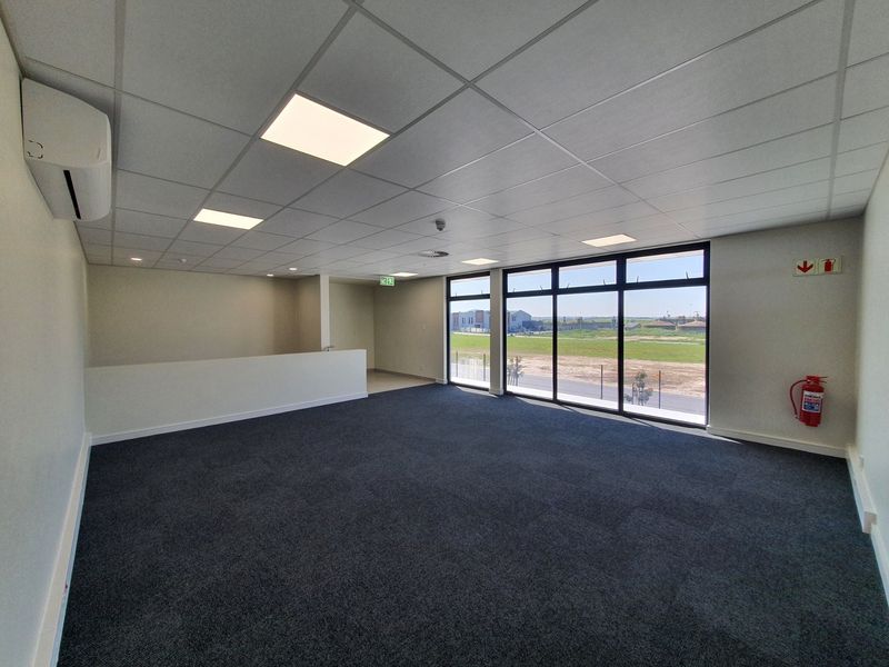 A modern, open-plan office space featuring carpeted flooring, a suspended ceiling with recessed lighting, split-system air conditioning, and large windows with exterior views.
