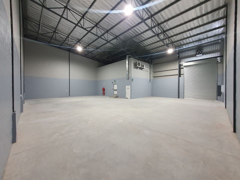 Spacious industrial warehouse unit featuring high ceilings with exposed steel trusses, concrete flooring, and a large roller shutter door for easy access.